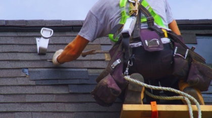 A roofer in a safety harness repairs shingles on a roof with tools, secured by a rope. The sky is partly cloudy.