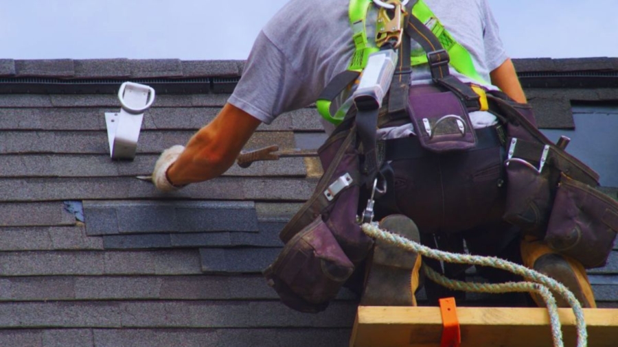 A roofer in a safety harness repairs shingles on a roof with tools, secured by a rope. The sky is partly cloudy.