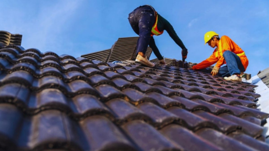 Two roofing contractors in safety gear installing dark ceramic roof tiles under a clear blue sky – "roofing near me" concept.
