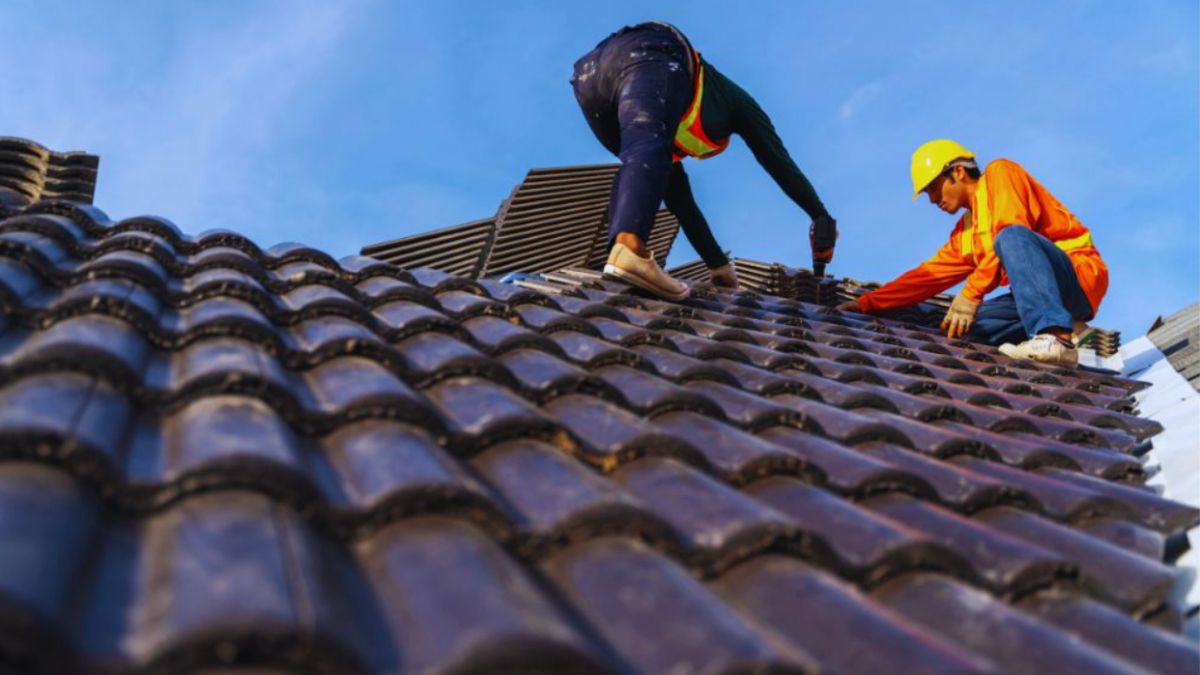 Two roofing contractors in safety gear installing dark ceramic roof tiles under a clear blue sky – "roofing near me" concept.