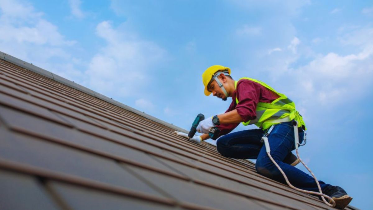 Roofer in safety gear and a yellow hard hat using a power drill on a sloped metal roof under a blue sky – concept of professional "roofing near me" services.