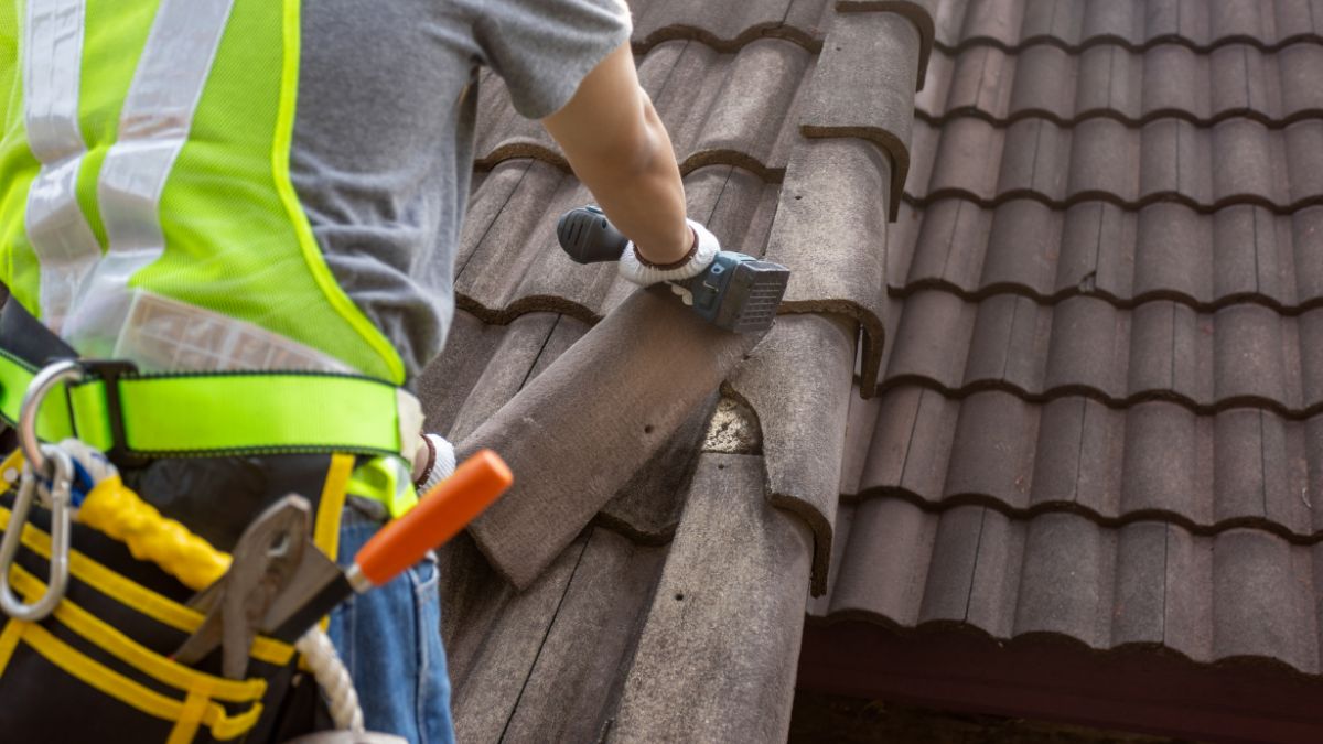 A construction worker wearing a safety harness and reflective vest repairs a clay tile roof, securing a ridge cap tile with a specialized roofing tool.