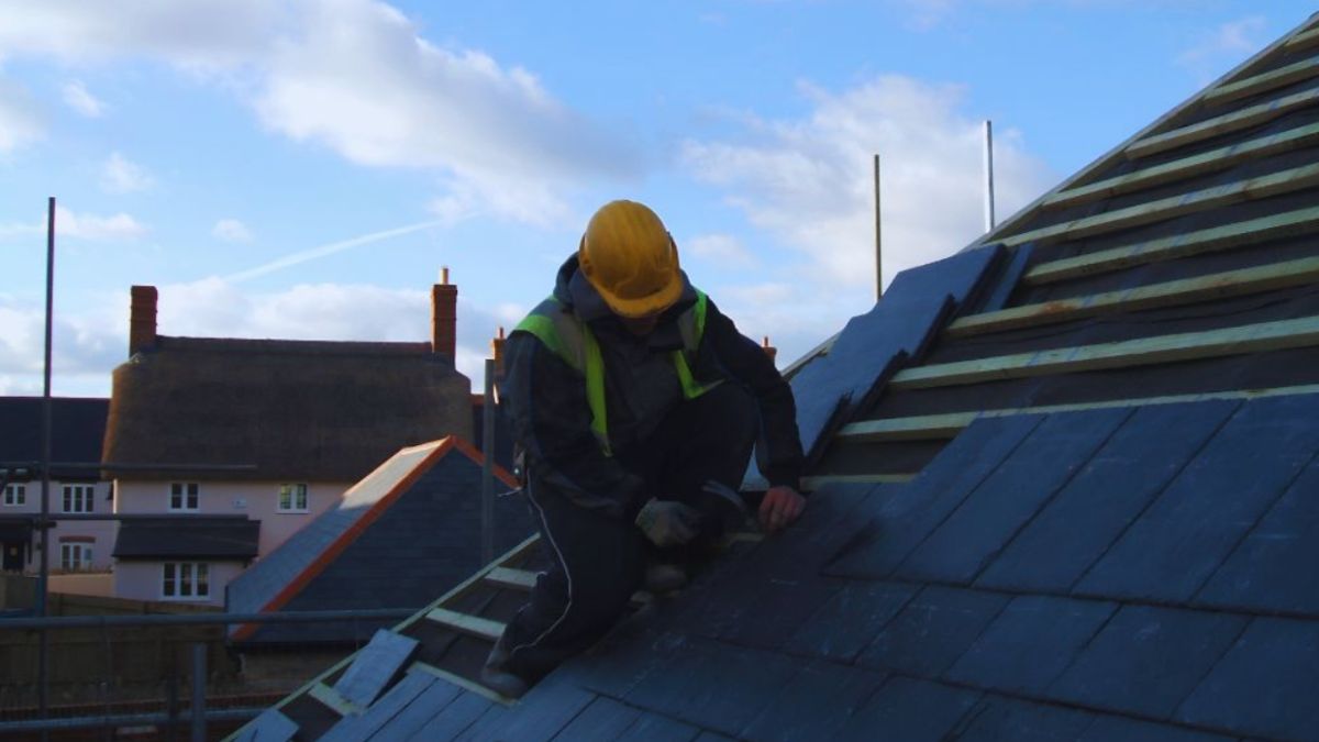 A roofer wearing a hard hat and safety vest is working on a residential roof repair.