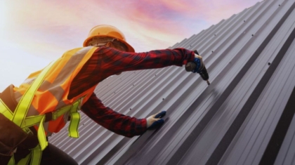 A roofing contractor wearing a hard hat, high-visibility safety vest, and gloves uses a power drill to secure a metal roof under a colorful sunset sky. The scene highlights professional roofing work in South Carolina.