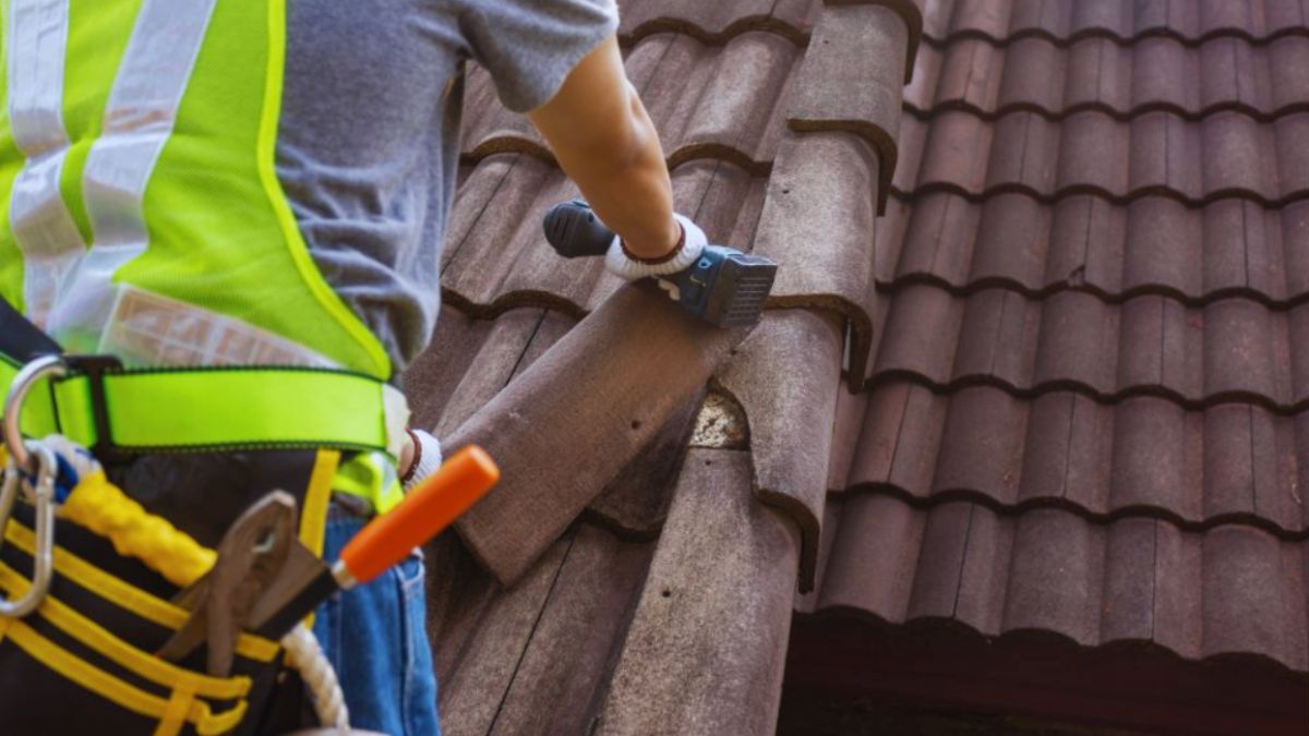 A roofing professional wearing a safety harness and vest uses a tool to inspect or repair brown ceramic roof tiles. The worker is focused on the ridge area, ensuring the tiles are properly aligned and secured after possible storm damage.