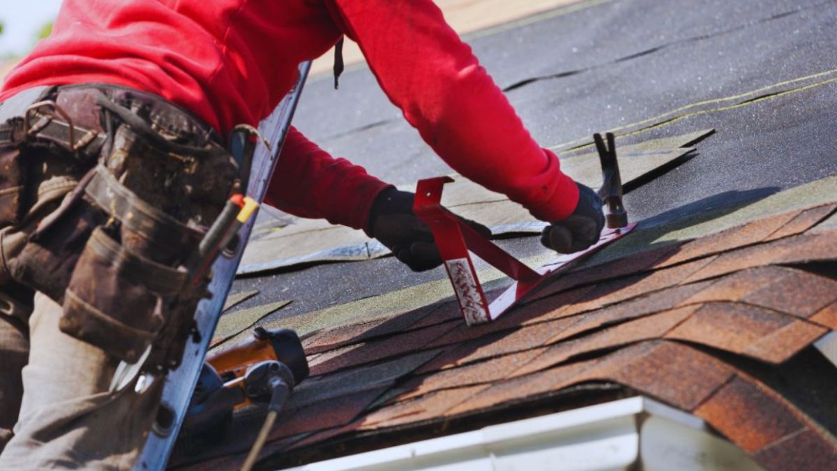 A worker wearing a red shirt and a tool belt is repairing a shingle roof; they are using technical tools to secure new shingles on a house roof, with roofing felt and equipment visible around the workspace.