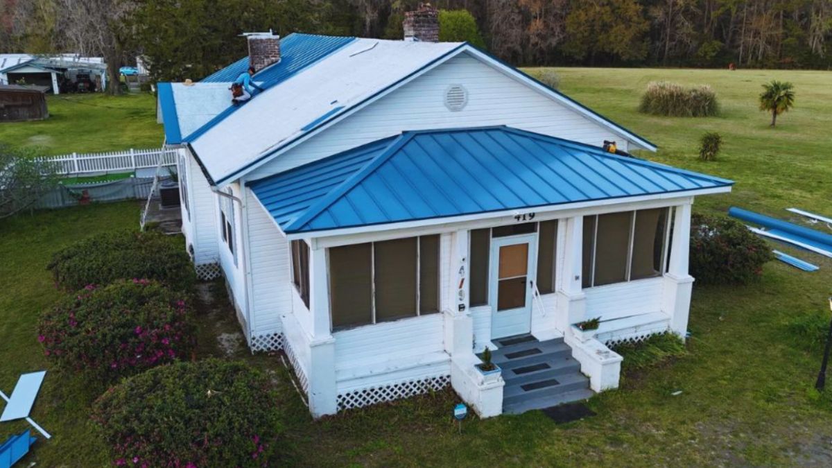 Professional emergency roof repair crew restoring a blue metal roof on a white farmhouse-style home in Bluffton, SC after storm damage, with safety equipment and materials visible around the property.