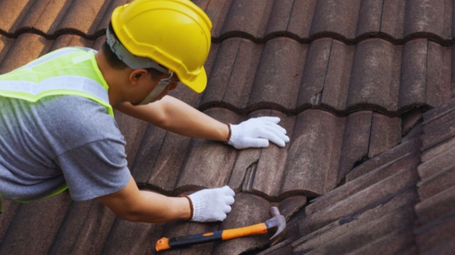 Construction worker in safety gear performing emergency roof repair on damaged brown tile shingles with a hammer on a residential home in Bluffton, SC.