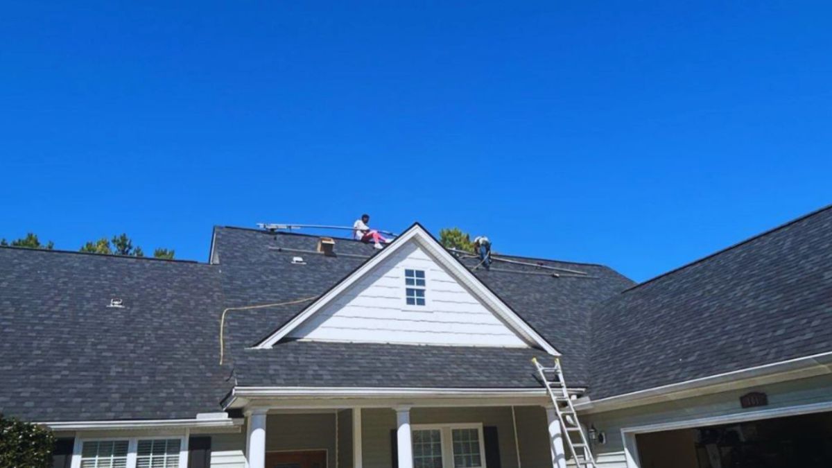 Roofing contractors repairing a roof leak on a single-story Bluffton, SC home, with workers on the dark-shingled roof above the front porch, ladder access, tools on the driveway, and a black convertible parked in front.