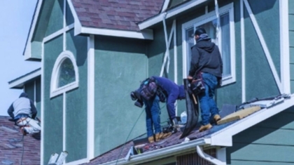 Roofers repairing a residential roof leak on a green two-story house in Bluffton, SC, using safety harnesses and tools while standing on the shingles near an upper-story window.