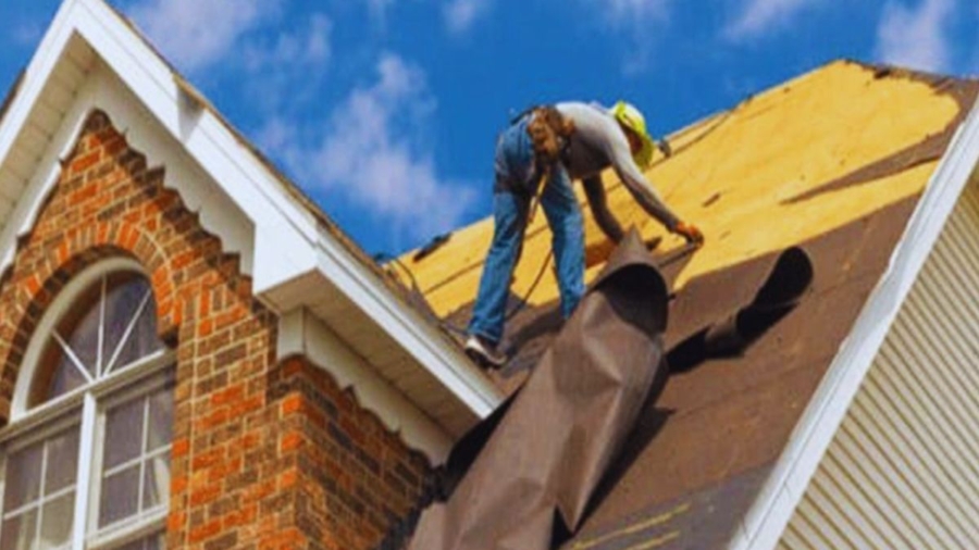 Roof replacement Ridgeland project showing a worker installing new roofing underlayment on a steep house roof, with exposed plywood decking and a brick gable featuring an arched window against a bright blue sky.