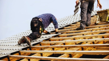 Workers installing metal roofing panels on a wooden roof frame, one kneeling and one standing with tools and materials, illustrating professional roofing services by Bluffton Roofing Company.