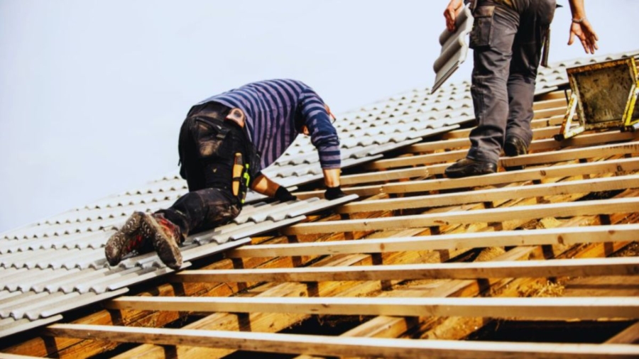 Workers installing metal roofing panels on a wooden roof frame, one kneeling and one standing with tools and materials, illustrating professional roofing services by Bluffton Roofing Company.