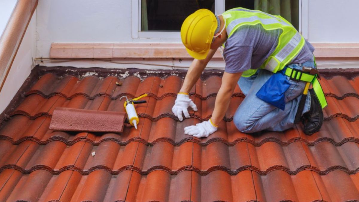 Roofing contractor in a hard hat and safety vest kneeling on a red tile roof, inspecting and repairing tiles near a window, representing a trusted roofing company in Bluffton, SC.