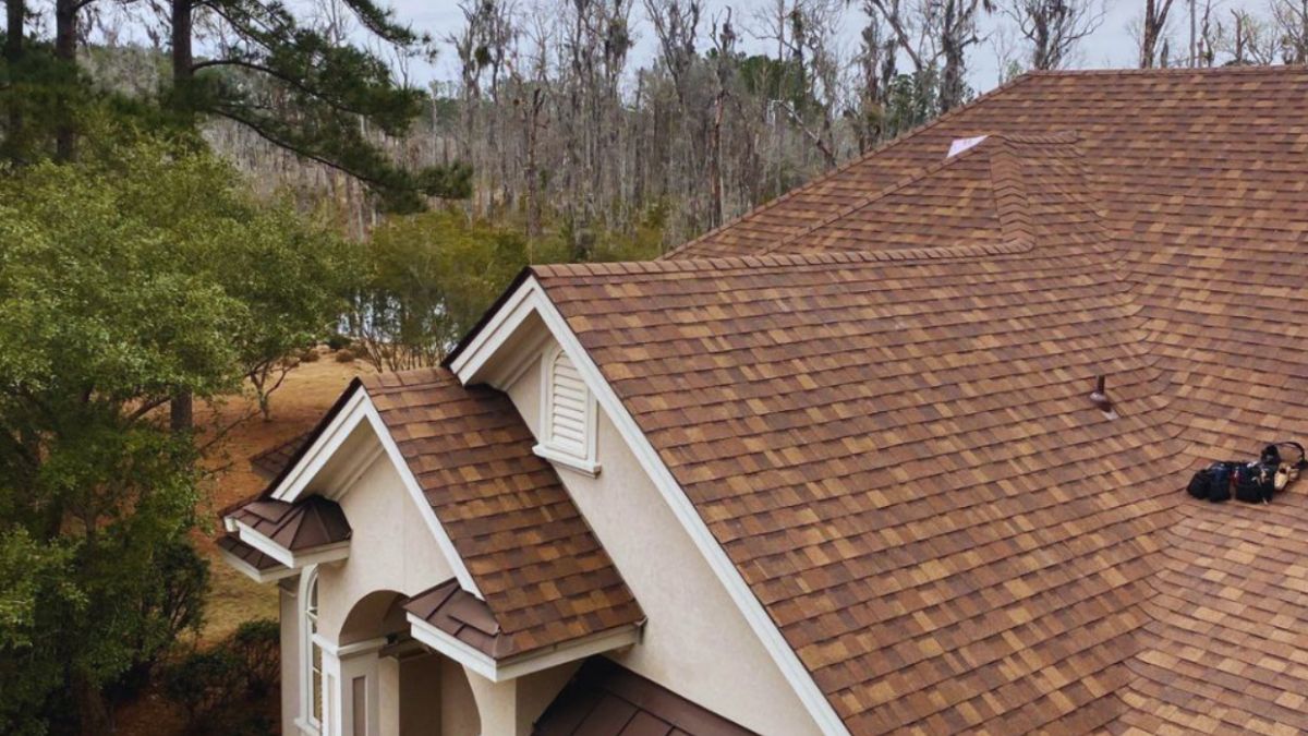 Aerial view of a tan stucco home in Hilton Head, SC, with a large brown shingle roof recently repaired, surrounded by tall trees and landscaping under an overcast sky.