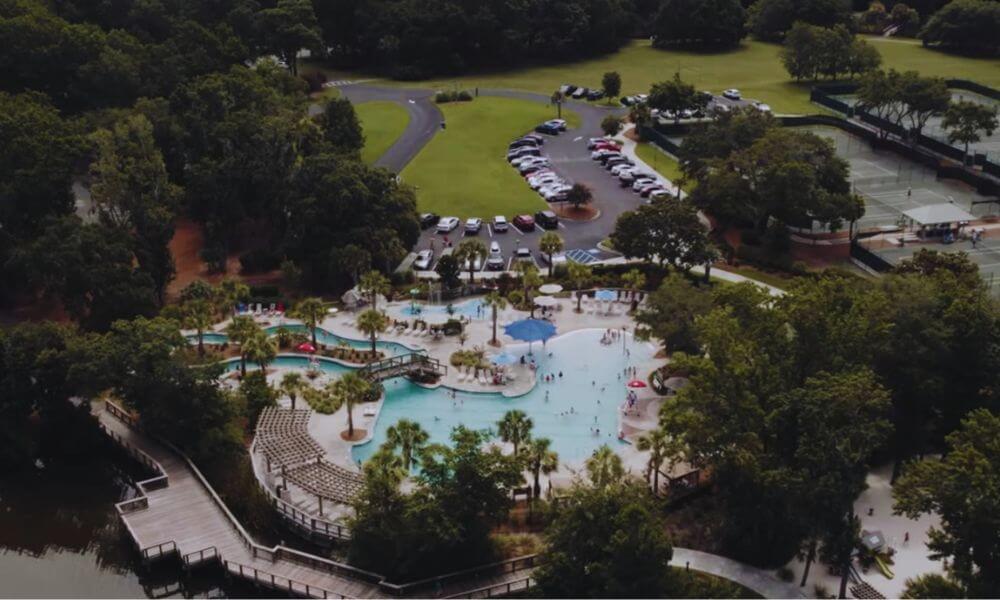 Aerial view of a resort-style pool complex surrounded by trees, boardwalks, tennis courts, and parking in Hilton Head Plantation on Hilton Head Island.