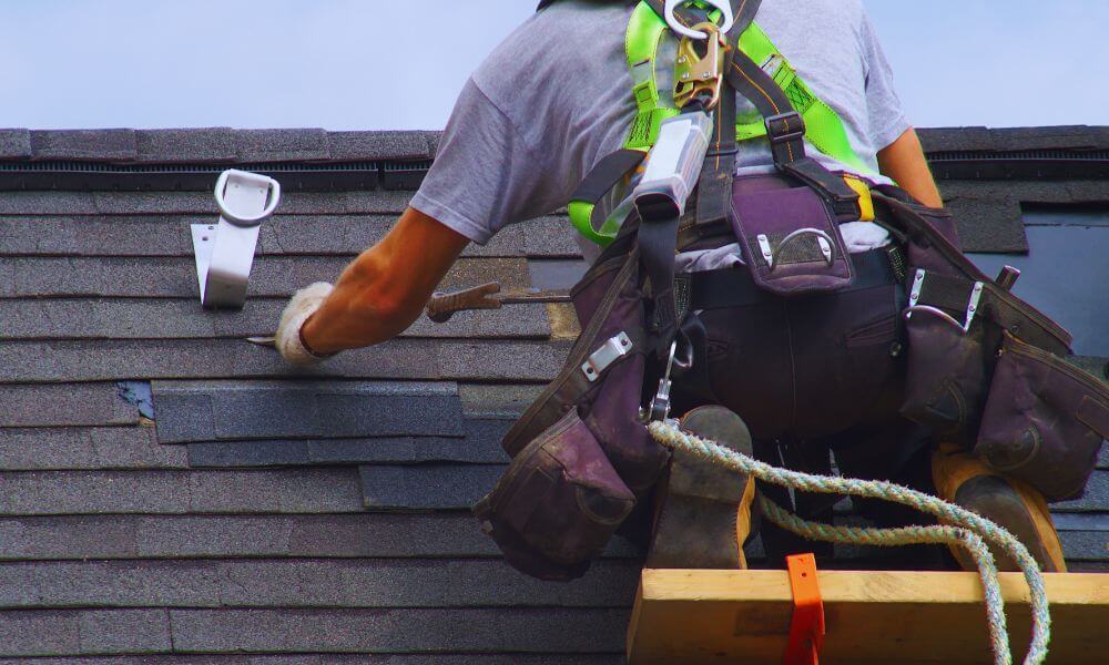Professional roofer in a safety harness repairing asphalt shingles on a steep roof, representing professional roofing services in Hilton Head Plantation.