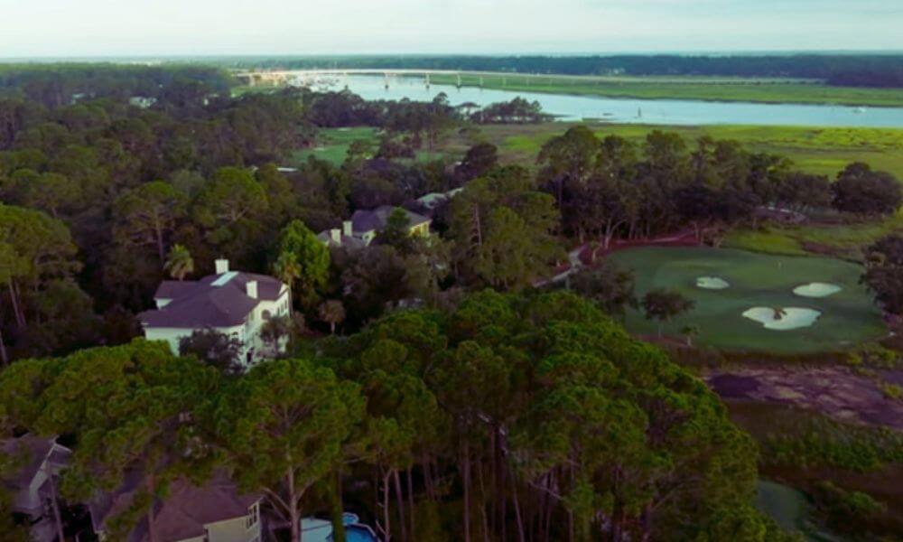 Aerial view of homes and tall pine trees in the Long Cove community on Hilton Head Island, overlooking a golf course and the waterway in the distance.