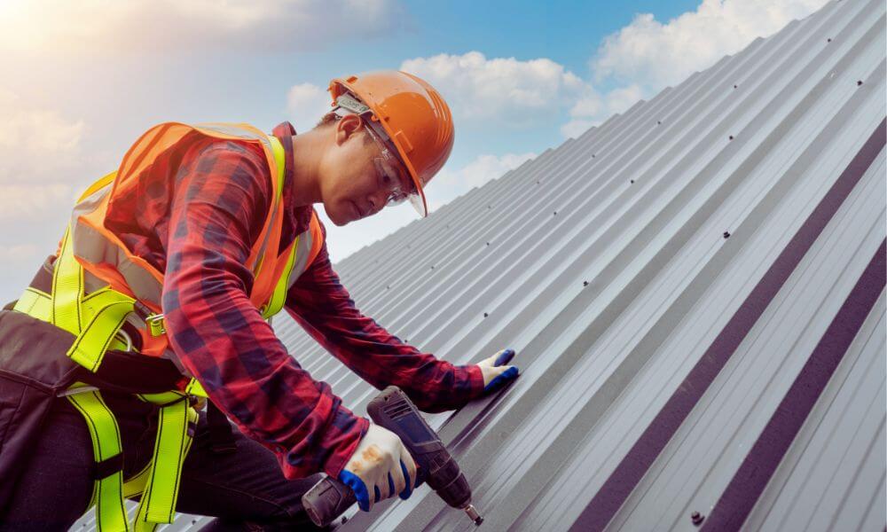 Professional roofer in safety harness and hard hat using a power drill to install fasteners on a sloped metal roof under a blue sky.