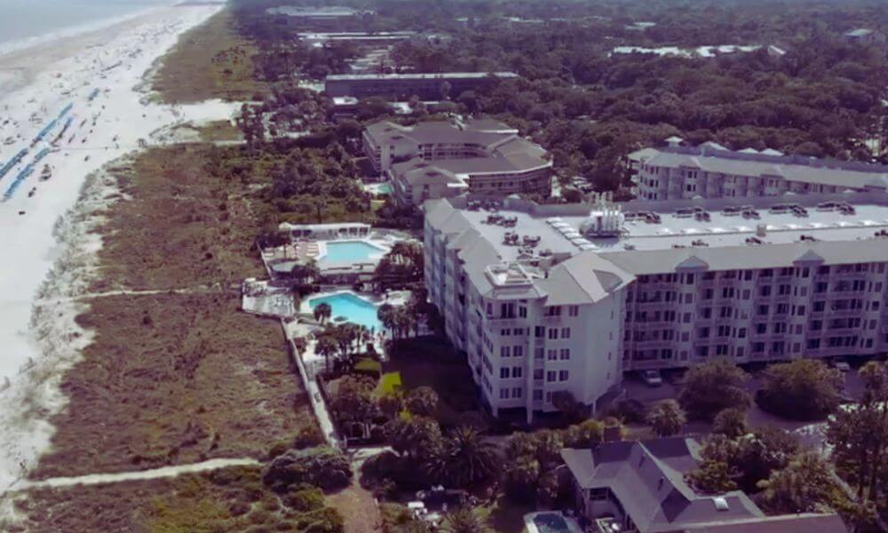 Aerial view of South Forest Beach in Hilton Head Island, SC, showing beachfront condos, resort buildings, swimming pools, and the sandy shoreline along the Atlantic coast.