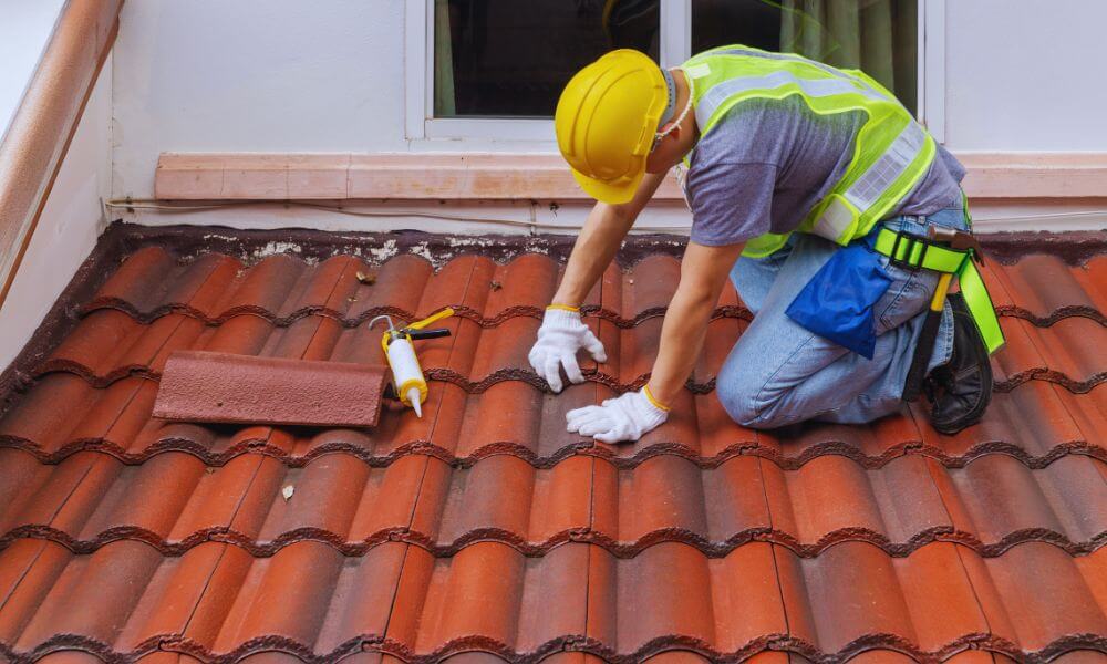 Professional roofer performing roofing services on a tile roof, repairing and installing roof tiles while wearing safety gear and using roofing tools.
