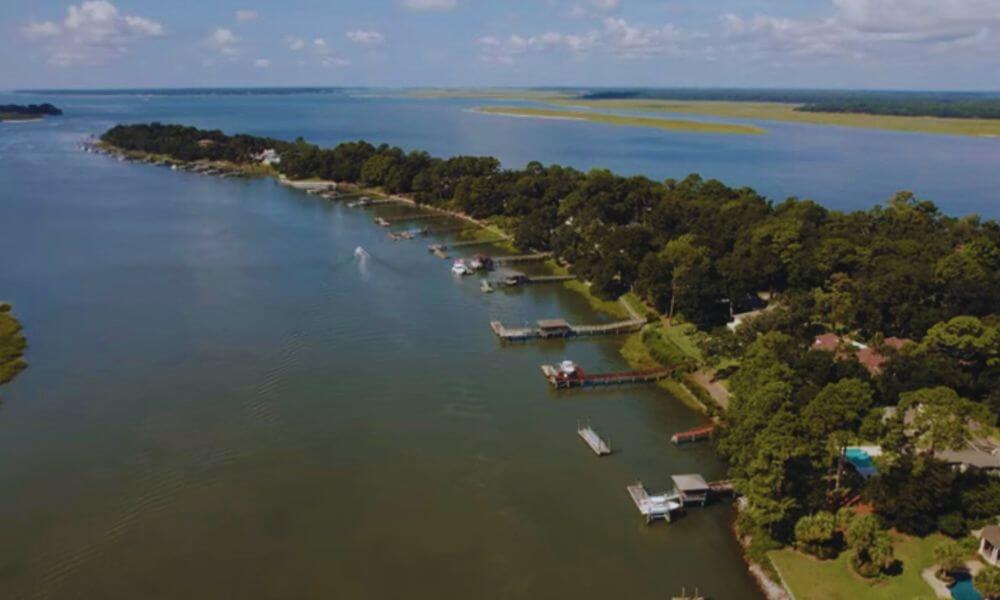 Aerial view of Spanish Wells in Hilton Head, SC, showing waterfront homes, private docks, lush trees, and calm coastal waterways under a blue sky.