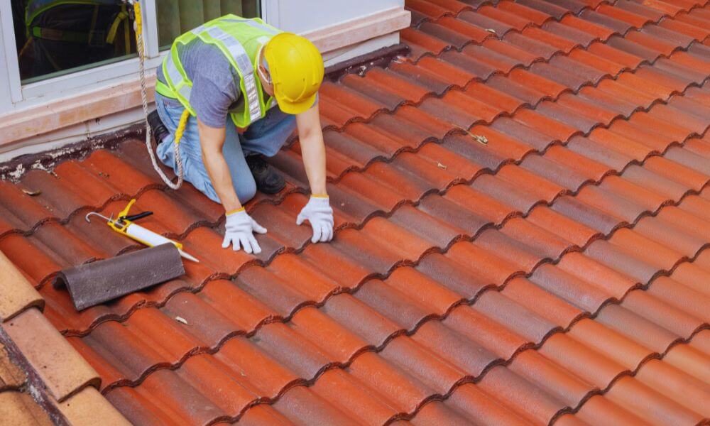 Roofing contractor in safety gear inspecting and repairing red clay tile roof near a window, representing professional roofing services.