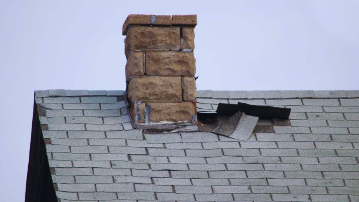 Damaged asphalt shingle roof with missing and lifted shingles around a masonry chimney.