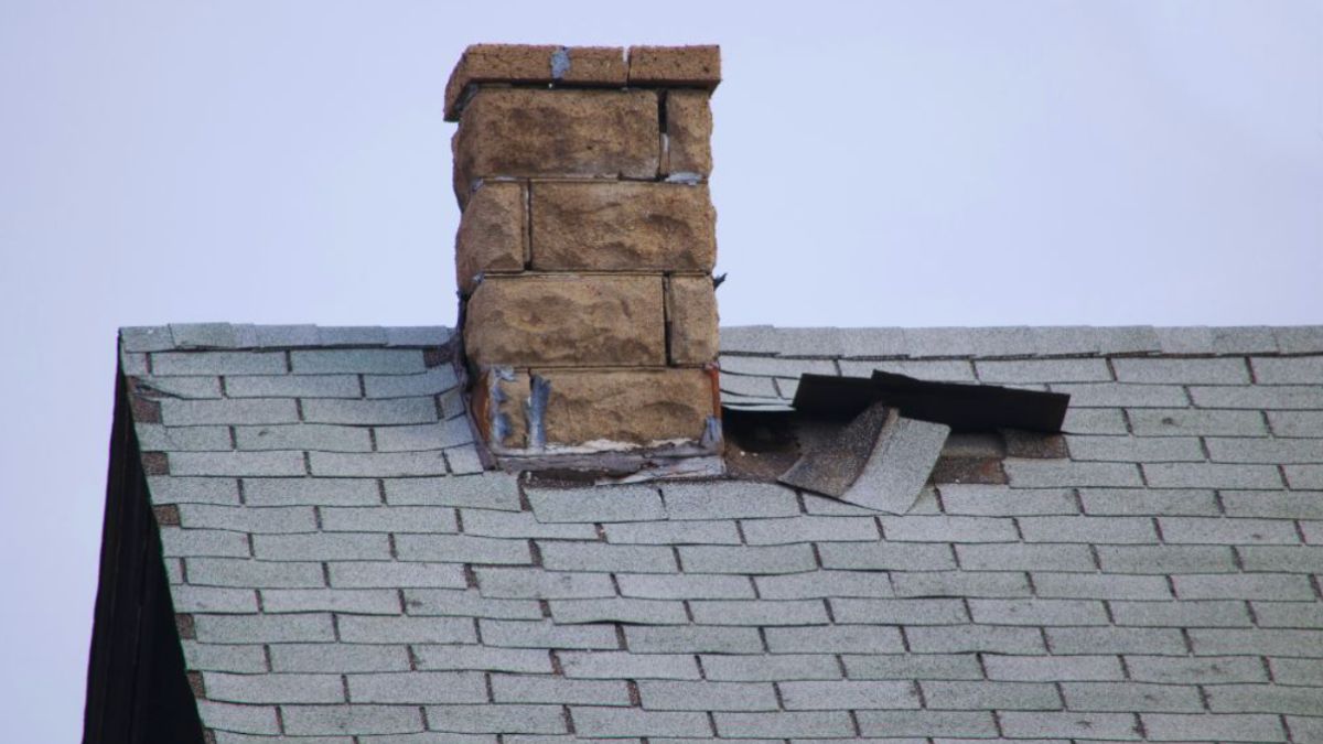 Damaged shingle roof with missing and lifted shingles near a brick chimney, showing exposed underlayment and storm-related roof damage.