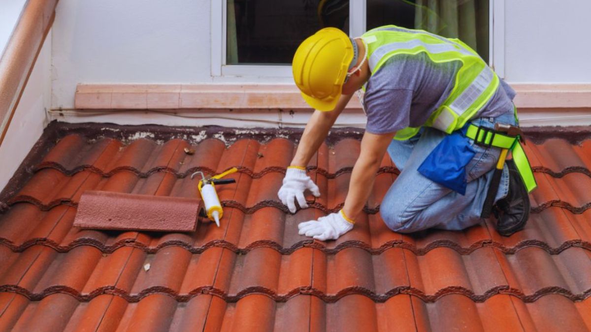 Roofing contractor in safety gear kneeling on red tile roof, sealing a damaged section during emergency roof repair in Hilton Head.