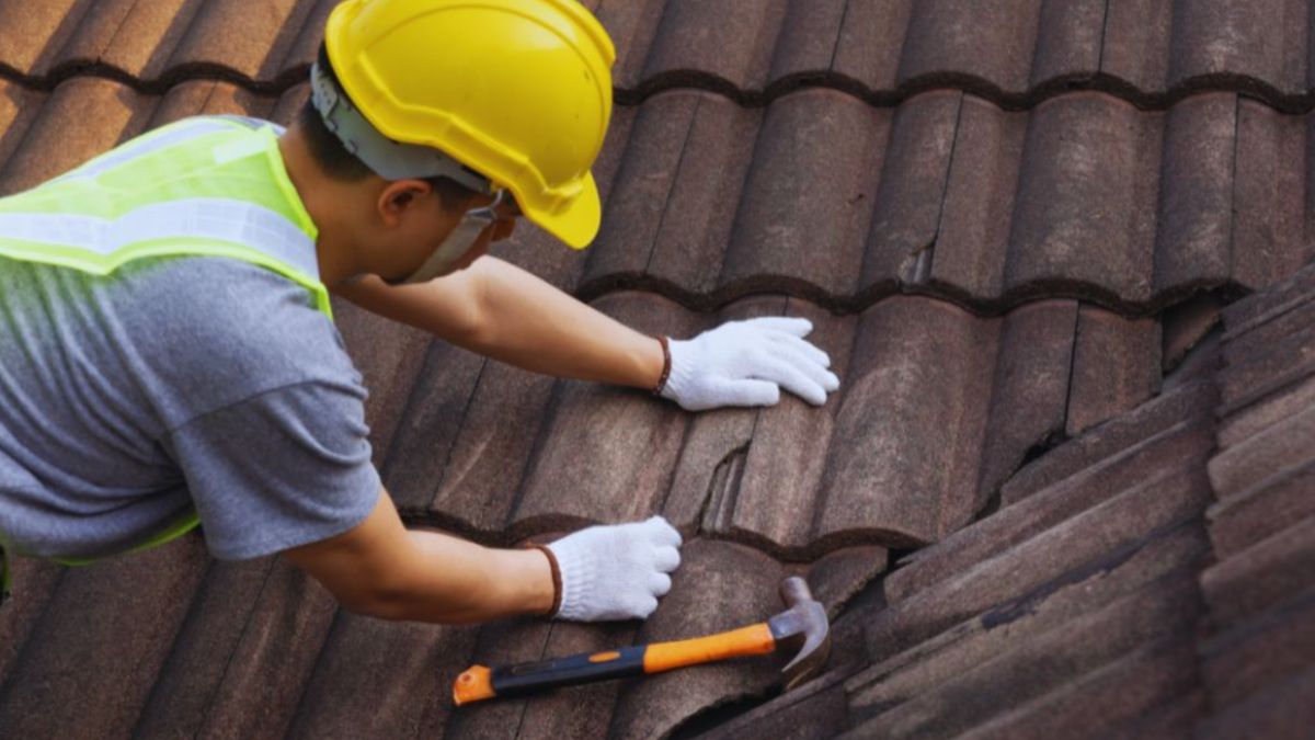 Roofing contractor repairing damaged brown tile roof with safety gear and tools, providing professional roofing services.
