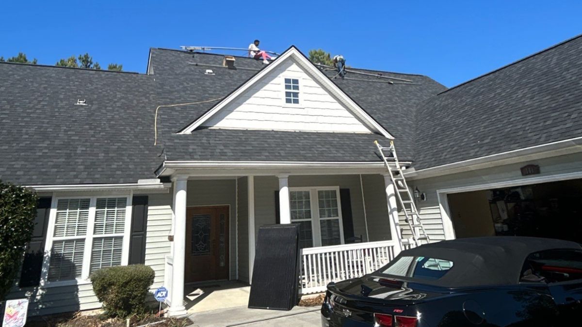 Roofing crew working on a dark shingle roof of a residential home near Rephaim Plantation in Bluffton, SC, with ladders and equipment visible in the driveway during residential roofing services.