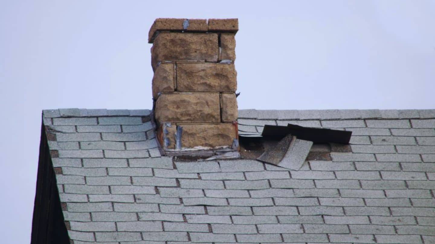 Damaged asphalt shingle roof with missing shingles and exposed roofing near a chimney.